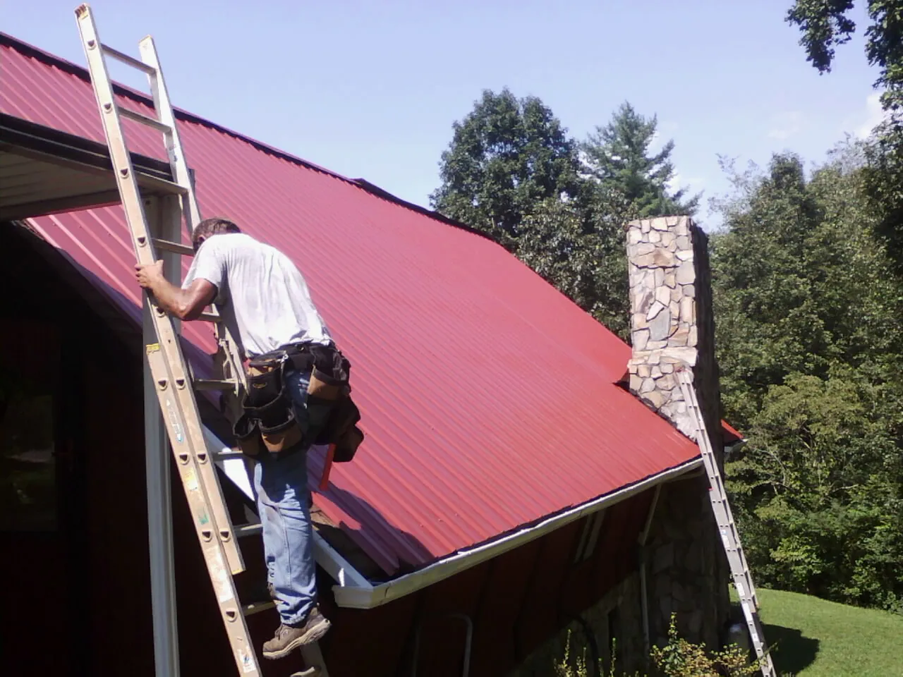 Roofer on a ladder beside a red metal roof and stone chimney — Soffit & Fascia Repair by Roofing Brothers in Granite City